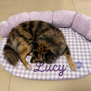 May include: A plush pet bed with a light purple gingham pattern and a soft, raised bolster edge. The center features the name Lucy embroidered in dark purple script, alongside a small heart-shaped patch with the text UPY.