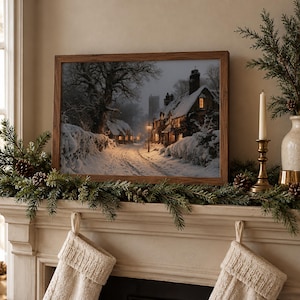 May include: A framed landscape print depicting a snow-covered village street at twilight. The scene features stone cottages with glowing windows, a tall chimney, and a street lamp illuminating a path through deep snow and trees.