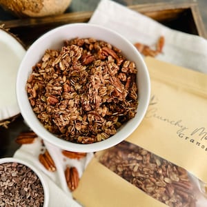 May include: A white bowl filled with chocolate granola with pecans. The granola is in a brown paper bag with the text "Crunchy Mom Granola".