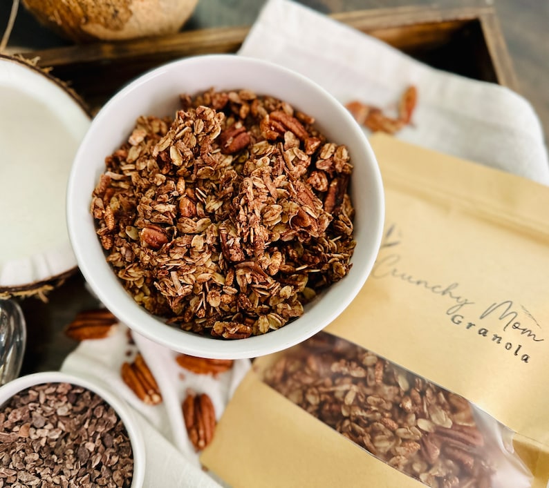 May include: A white bowl filled with chocolate granola with pecans. The granola is in a brown paper bag with the text "Crunchy Mom Granola".