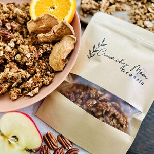 May include: A brown paper bag with a clear window showing granola inside. The bag has a logo that says "Crunchy Mom Granola". There are dried apple slices, pecans, and an orange slice in the background.