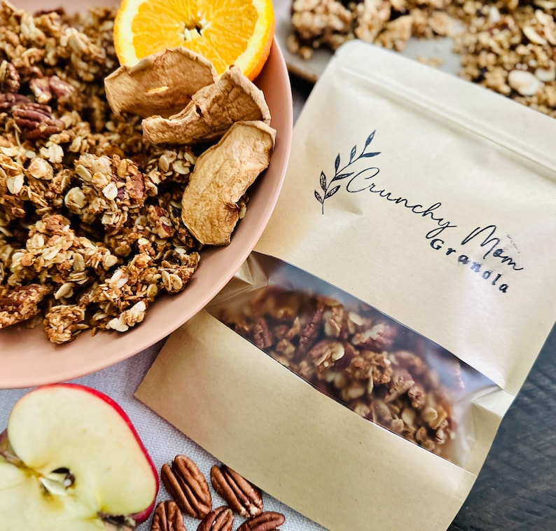 May include: A brown paper bag with a clear window showing granola inside. The bag has a logo that says "Crunchy Mom Granola". There are dried apple slices, pecans, and an orange slice in the background.