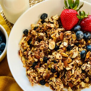 May include: A white bowl filled with granola, blueberries, and sliced strawberries. The granola is a mixture of oats, nuts, and seeds. The bowl is on a white plate with a yellow napkin in the background.