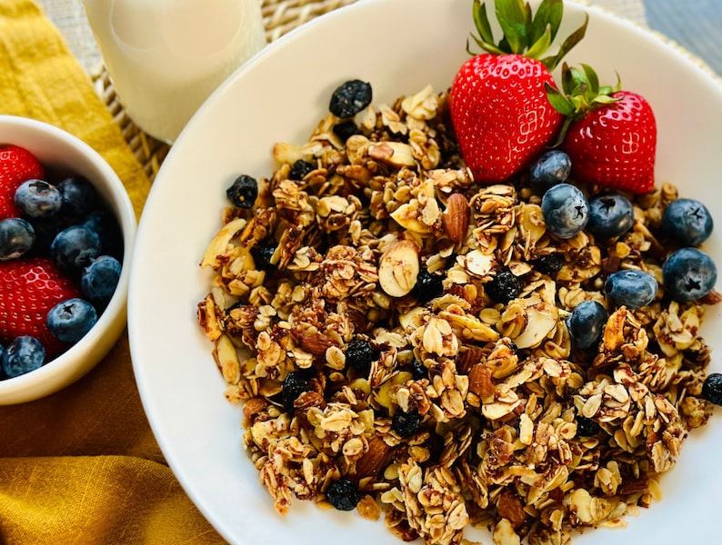 May include: A white bowl filled with granola, blueberries, and sliced strawberries. The granola is a mixture of oats, nuts, and seeds. The bowl is on a white plate with a yellow napkin in the background.