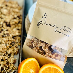May include: A brown paper bag with a clear window showing granola inside. The bag has the text "Crunchy Mom Granola" printed on it. There are two orange slices in the foreground.