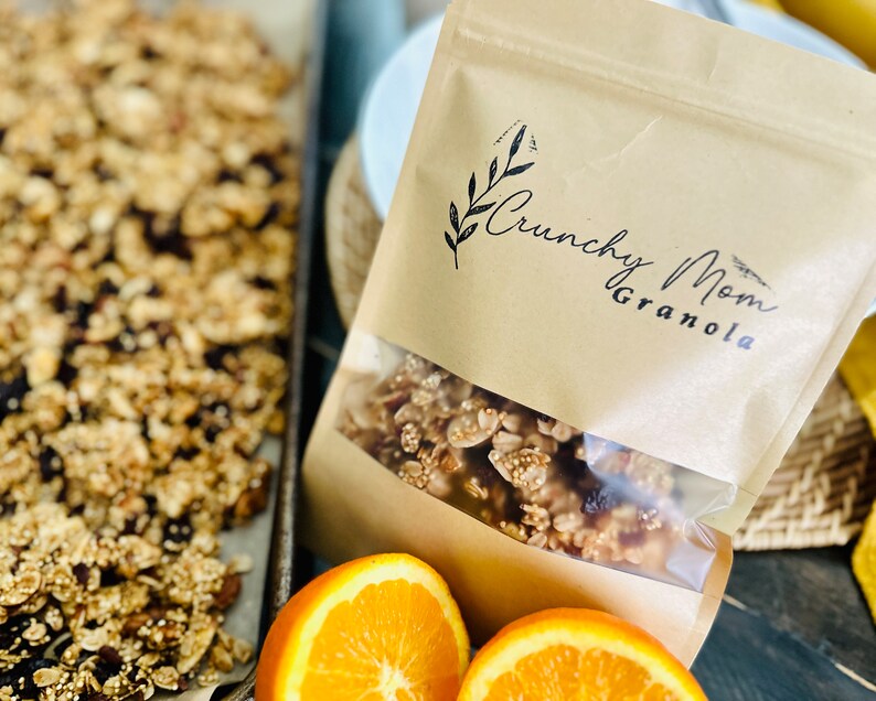 May include: A brown paper bag with a clear window showing granola inside. The bag has the text "Crunchy Mom Granola" printed on it. There are two orange slices in the foreground.