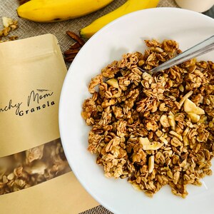 May include: A white bowl filled with crunchy granola with banana slices and nuts. The granola is labelled "Crunchy Mom Granola" on a brown paper bag.