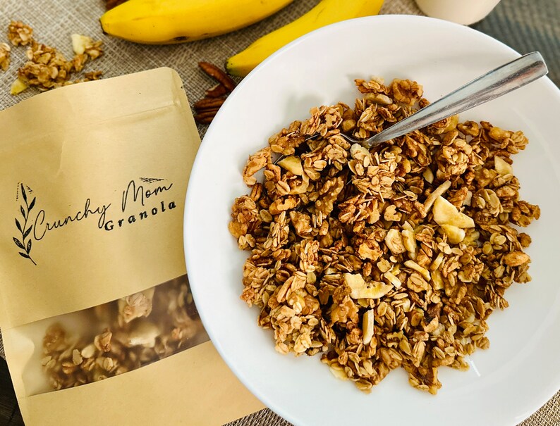 May include: A white bowl filled with crunchy granola with banana slices and nuts. The granola is labelled "Crunchy Mom Granola" on a brown paper bag.