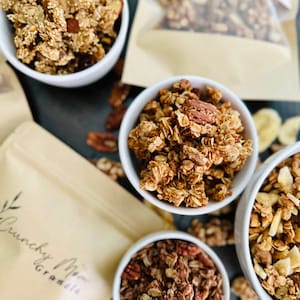 May include: Four white bowls filled with different types of granola. The granola is a mix of oats, nuts, and dried fruit. The bowls are on a dark surface with a brown paper bag in the background. The bag has the text "Crunchy Nut Granola" printed on it.