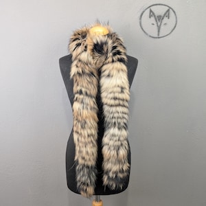 May include: A faux fur scarf in shades of brown, black, and cream, displayed on a black mannequin. The scarf has a textured, striped pattern and a soft appearance. The background is a plain gray wall.