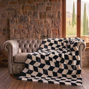 May include: A tufted leather sofa in a muted brown tone features a plush throw blanket draped over its arm and seat. The blanket displays a high-contrast black and white geometric diamond pattern, creating a bold visual texture against the furniture.