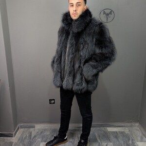 May include: A gray fur coat with a high collar and a zipper closure. The coat is worn with black pants and black sneakers with brown soles. The background is a plain gray wall.