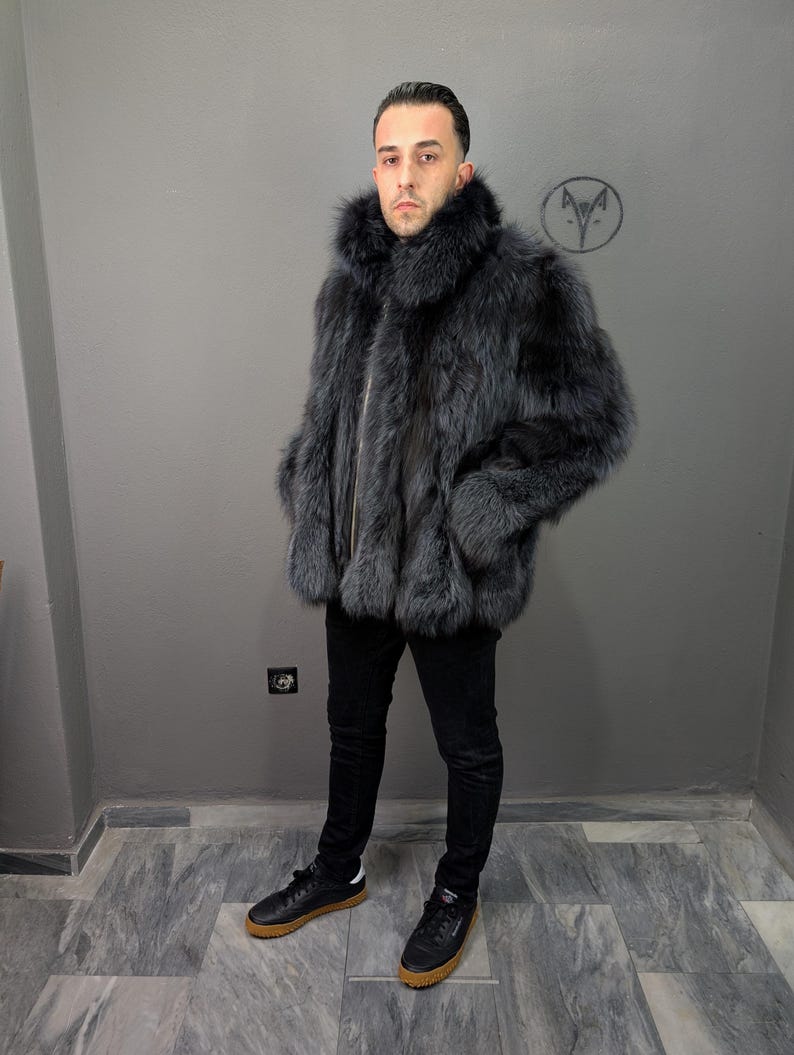 May include: A gray fur coat with a high collar and a zipper closure. The coat is worn with black pants and black sneakers with brown soles. The background is a plain gray wall.