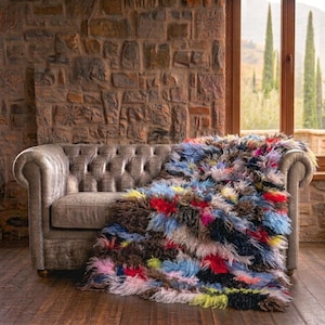 May include: A tufted, long-pile throw blanket featuring a vibrant, multicolored patchwork design with shades of red, blue, yellow, pink, and black. The shaggy, textured textile is draped over a tufted, gray leather chesterfield sofa.