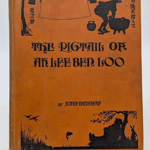 以下が含まれることがあります： ジョン・ベネット著「The Pigtail of Ah Lee Ben Loo」というタイトルの古書。オレンジ色の表紙には、人物、鍋、物干し台の黒いイラストが描かれており、日本語のテキストも含まれています。