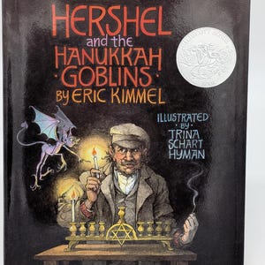 May include: A hardcover book titled "Hershel and the Hanukkah Goblins" by Eric Kimmel. The cover features an illustration of a man lighting candles on a menorah, with a goblin and text. A silver "Caldecott Honor Book" seal is on the cover.