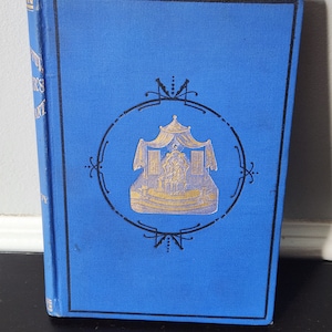 Può includere: Un libro rilegato in blu con un'illustrazione dorata e nera di un edificio con un tetto a punta e una corona in cima. Il titolo del libro è "The Adventures of", ma il resto del titolo è oscurato.