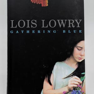 May include: A hardcover book titled "Gathering Blue" by Lois Lowry. The cover features a young person with long dark hair, holding flowers. The background is black, with the title in large, light gray letters.