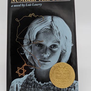 May include: A hardcover book titled "NUMBER THE STARS" by Lois Lowry. The cover features a black background with a portrait of a young girl and a gold Star of David necklace. A gold medal is affixed to the cover.