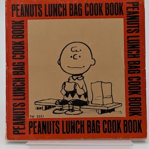 May include: A vintage "Peanuts Lunch Bag Cook Book" featuring Charlie Brown on the cover. The book has a brown background with the title in red. Charlie Brown is holding a sandwich. The book is a square shape.