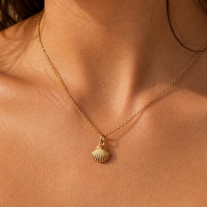 May include: A gold seashell pendant necklace. The necklace has a delicate chain and a small, detailed seashell charm. The pendant hangs from a thin chain. The person is wearing a white vest top. The necklace is a piece of jewellery.