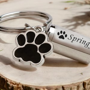 May include: A silver pet memorial keychain with a black paw print charm and a small cylinder engraved with "Springy" and a paw print. The keychain is on a wooden surface with greenery.