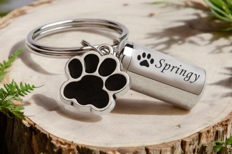 May include: A silver pet memorial keychain with a black paw print charm and a small cylinder engraved with "Springy" and a paw print. The keychain is on a wooden surface with greenery.