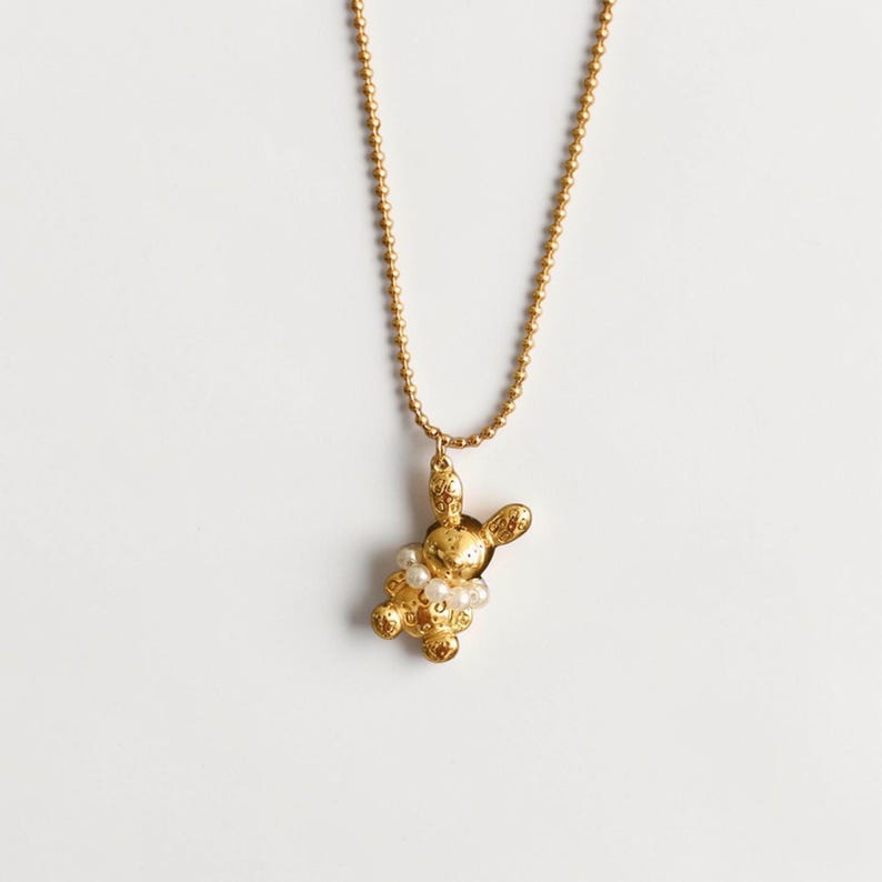 May include: A gold-colored bunny pendant necklace with a beaded chain. The bunny pendant is adorned with small white pearls around its neck. The necklace is displayed against a white background.