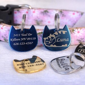 May include: A pink floral pet collar with five cat-shaped metal tags in blue, gold, silver, and black. The tags feature engraved text including addresses, phone numbers, and names like Luna and Bella, each attached with a circular metal ring.