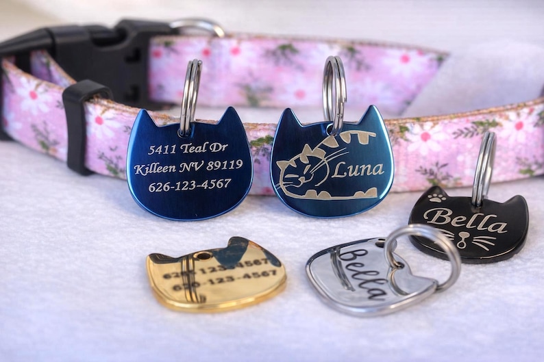 May include: A pink floral pet collar with five cat-shaped metal tags in blue, gold, silver, and black. The tags feature engraved text including addresses, phone numbers, and names like Luna and Bella, each attached with a circular metal ring.