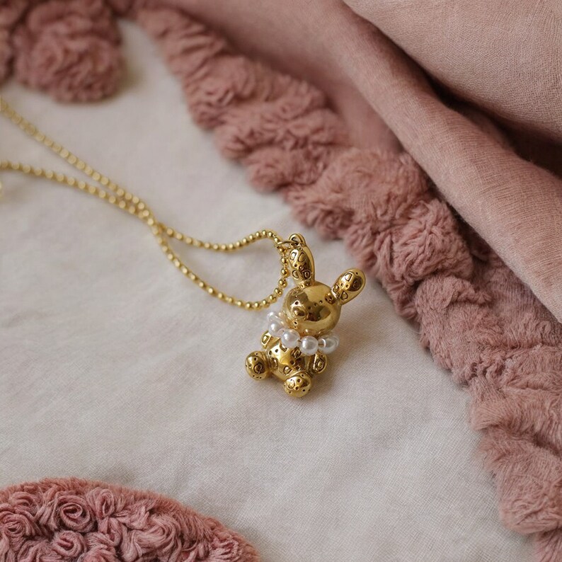 May include: A gold-colored bunny pendant necklace with a pearl collar. The bunny has a textured surface and is suspended from a delicate gold chain. The background features soft pink fabric.