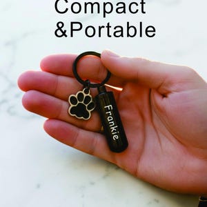 May include: Black keychain with a paw print charm and a small, black cylinder engraved with the name "Frankie." The text "Compact & Portable" is above the keychain. The keychain is held in a hand.