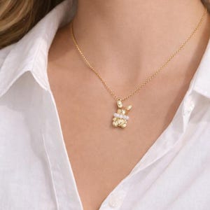 May include: A gold-colored necklace featuring a pendant shaped like a bunny. The bunny pendant has a textured surface and is adorned with small pearls and clear gemstones. The necklace is worn against a white shirt.
