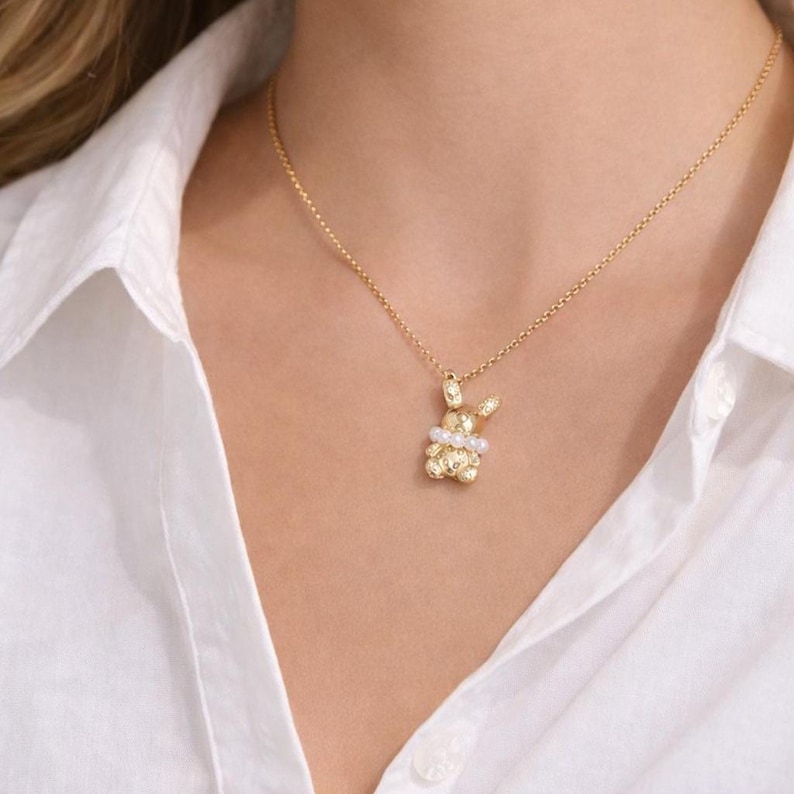 May include: A gold-colored necklace featuring a pendant shaped like a bunny. The bunny pendant has a textured surface and is adorned with small pearls and clear gemstones. The necklace is worn against a white shirt.