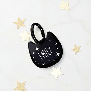 May include: A black cat-shaped pet tag featuring the name EMILY in white capital letters, surrounded by small white star icons. The tag includes a black metal split ring attached to the top for secure fastening to a collar or accessory.