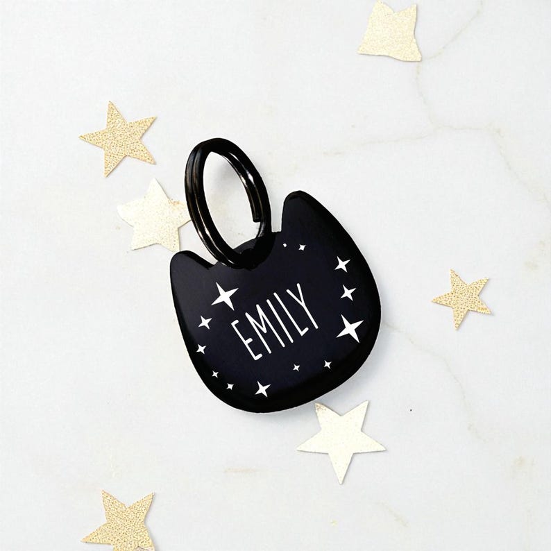 May include: A black cat-shaped pet tag featuring the name EMILY in white capital letters, surrounded by small white star icons. The tag includes a black metal split ring attached to the top for secure fastening to a collar or accessory.