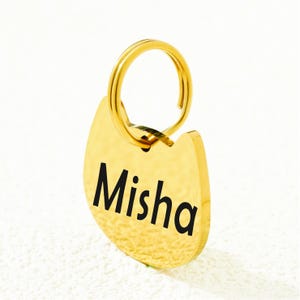 May include: A gold-colored metal pet ID tag shaped like a cat head with a circular split ring attached at the top. The tag features the name Misha engraved in a bold, black, sans-serif font across the center of the smooth, reflective surface.