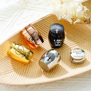 Puede incluir: Una colección de pequeñas urnas metálicas en tonos dorados, oro rosa y plateados. Algunas urnas presentan textos grabados, incluyendo nombres y fechas, junto con diseños de corazón y árbol. Una urna negra tiene una cruz y el texto "Sorry I had to split."