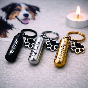 Puede incluir: Tres llaveros conmemorativos para mascotas en negro, plateado y dorado. Cada llavero tiene un colgante cilíndrico con un nombre y una huella de pata, y un dije en forma de pata. El fondo incluye una imagen de perro y una vela encendida.