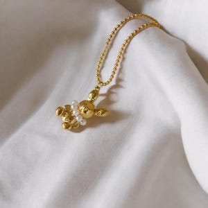 May include: A gold-colored necklace featuring a pendant shaped like a balloon animal. The pendant is adorned with small, white pearls and hangs from a delicate chain. The necklace is displayed on a white, textured fabric.