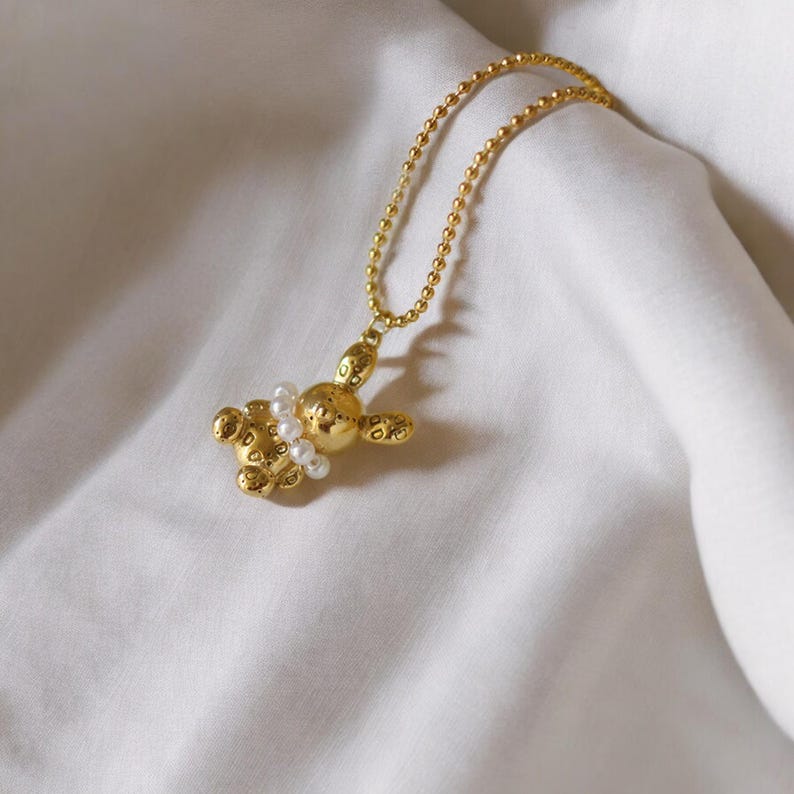 May include: A gold-colored necklace featuring a pendant shaped like a balloon animal. The pendant is adorned with small, white pearls and hangs from a delicate chain. The necklace is displayed on a white, textured fabric.