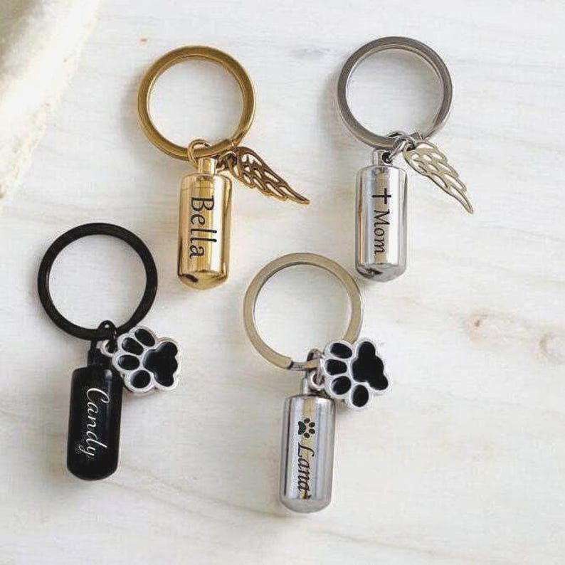 May include: Keychains with memorial pendants. One gold keychain has a pendant with the name "Bella" and a wing charm. Another silver keychain has a pendant with the name "Mom" and a wing charm. Two other keychains have paw print charms and the names "Candy" and "Lana".