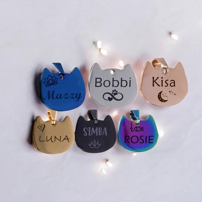 May include: Six cat-shaped pet ID tags in various colors and finishes. The tags are engraved with names and symbols: blue with Mazzy, silver with Bobbi, rose gold with Kisa, gold with Luna, black with Simba, and iridescent rainbow with Rosie.