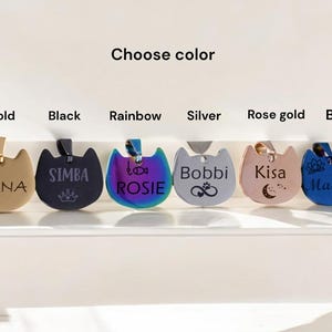 May include: Six cat-shaped pet tags in gold, black, rainbow, silver, rose gold, and blue. Each tag features custom engravings including names like LUNA, SIMBA, ROSIE, Bobbi, Kisa, and Mazzy, along with icons like a heart, crown, fish, paw, and moon.