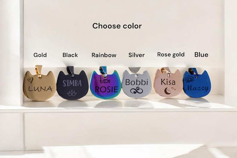 May include: Six cat-shaped pet tags in gold, black, rainbow, silver, rose gold, and blue. Each tag features custom engravings including names like LUNA, SIMBA, ROSIE, Bobbi, Kisa, and Mazzy, along with icons like a heart, crown, fish, paw, and moon.