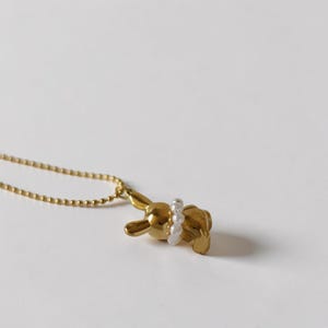 May include: A gold-colored bunny pendant necklace with a pearl accent. The bunny is lying down, and the pearls are around its waist. The necklace chain is a delicate gold color. The background is a plain white.