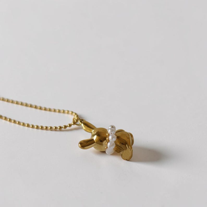 May include: A gold-colored bunny pendant necklace with a pearl accent. The bunny is lying down, and the pearls are around its waist. The necklace chain is a delicate gold color. The background is a plain white.