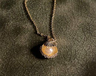 May include: A gold chain necklace with a seashell pendant. The seashell is decorated with small rhinestones and has a pearl inside.