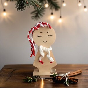 Puede incluir: Figura de madera con una cara sonriente, cabello trenzado rojo y blanco y una bufanda blanca. La figura está sobre una base de madera, con palitos de canela y vegetación en primer plano. Luces de hadas y ramas de hoja perenne en el fondo.