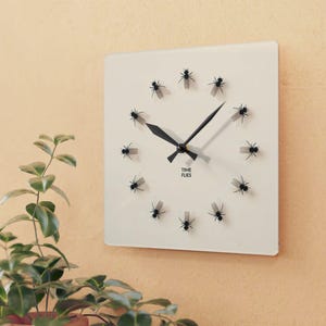 May include: Square wall clock with a cream-colored face. The clock features black hands and is decorated with small, black fly figures in place of numbers. The words "TIME FLIES" are printed below the clock's center.